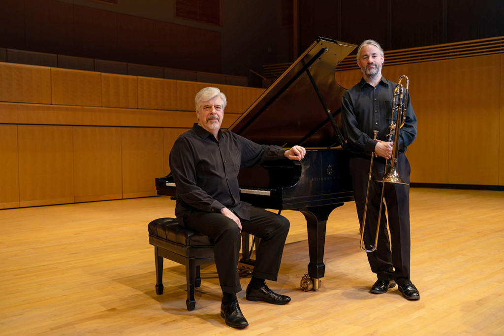 Two men, the one on the left at a piano, the one on the right holding a trombone.